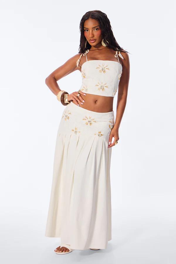 Woman modeling a cream cropped shirt, wide-leg pants, and a pink/yellow floral sarong with white lace. She wears sunglasses and gold heels.