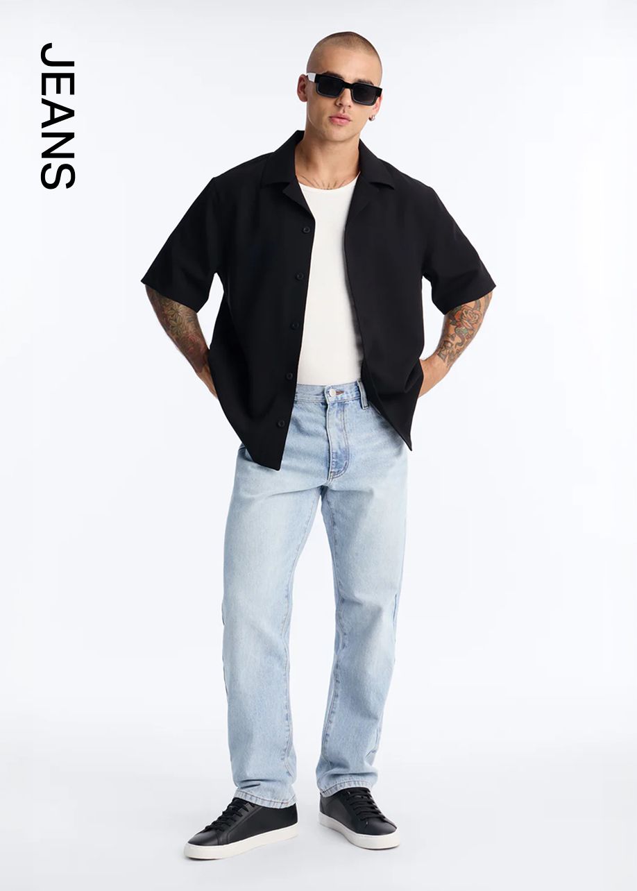 Man in black button-up shirt, white tee, light wash jeans, and black sneakers, hands on hips. Discover our new men's casual jeans.