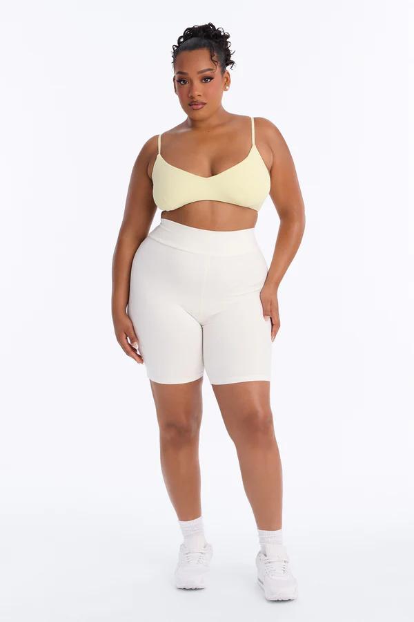 Confident plus-size woman with dark curly hair in a light yellow ribbed sports bra and cream high-waisted biker shorts, white sneakers, on a white background.