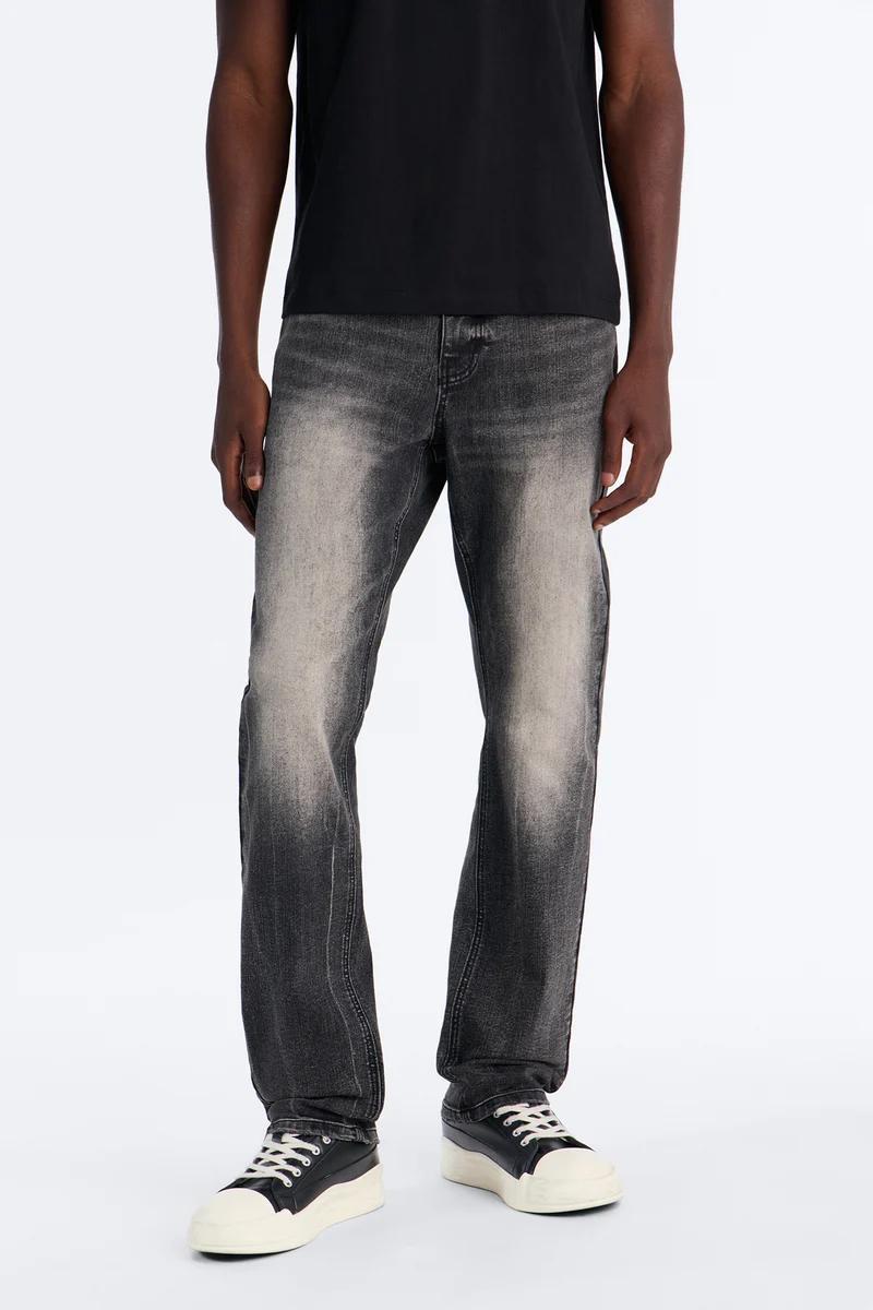 Model in black tee and faded gray straight-leg men's jeans with platform sneakers, highlighting modern denim fashion.