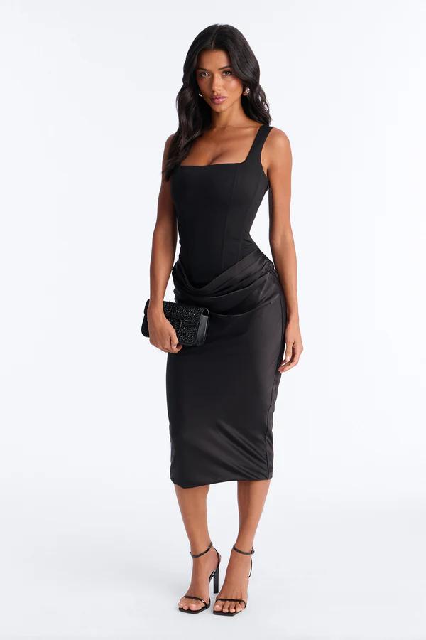Woman in a black square-neck midi dress with a corseted bodice and draped skirt, holding an embellished black clutch, wearing strappy heels.