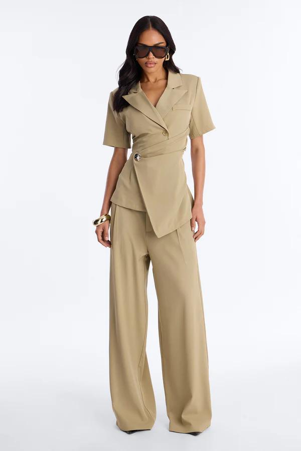 Woman in a cream sleeveless wrap top and matching wide-leg pleated pants, holding a small pouch bag and wearing flat sandals.