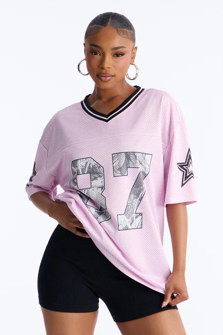 Woman in a light pink mesh "87" football jersey with black trim and a black star, wearing black ribbed shorts and silver hoop earrings.