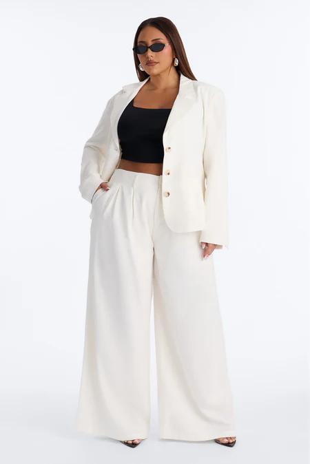 Mujer segura de cuerpo entero, vistiendo un blazer y pantalones anchos crema, un top corto negro y gafas de sol oscuras de ojo de gato.