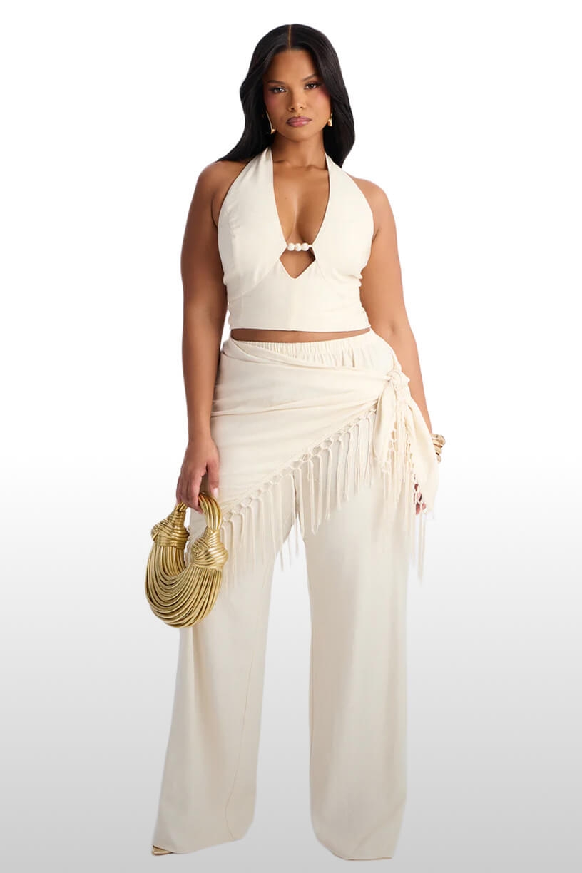 Woman in chic cream halter crop top, wide-leg pants with fringe sarong wrap, and gold bag. Perfect for plus size summer resort wear.
