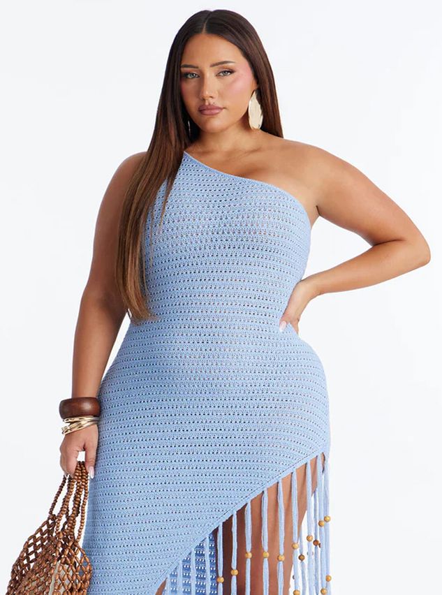 Woman modeling a light blue one-shoulder crochet dress with a beaded fringe hem, holding a woven bag. Ideal summer resort wear.