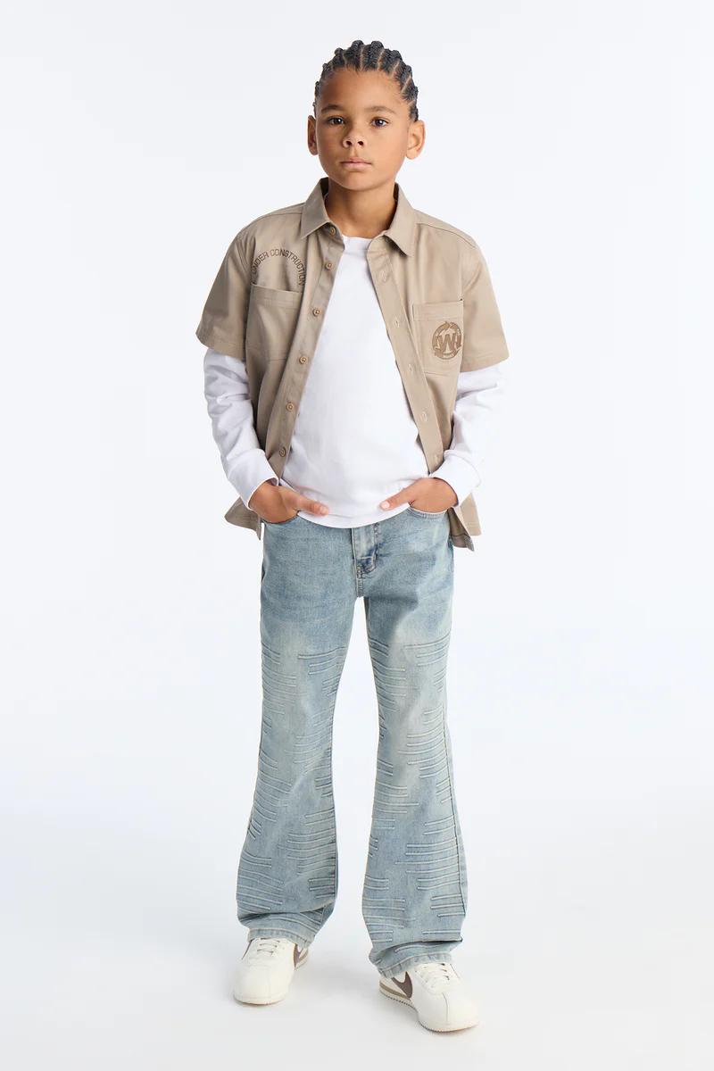 Niño en camisa utilitaria beige "UNDER CONSTRUCTION" sobre camiseta blanca, jeans acampanados celestes y zapatillas. Nueva moda casual infantil.