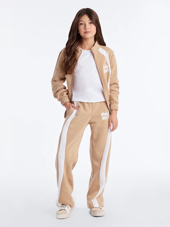 Young girl models a comfortable tan tracksuit with white side stripes and "Rich Girl" logo. Perfect girls' casual wear.