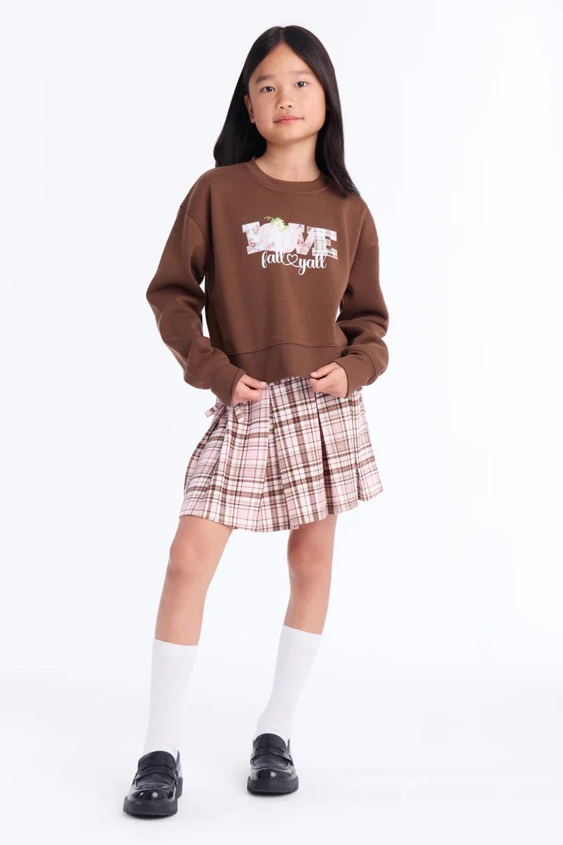 Niña con sudadera marrón y falda plisada a cuadros rosa con mocasines.