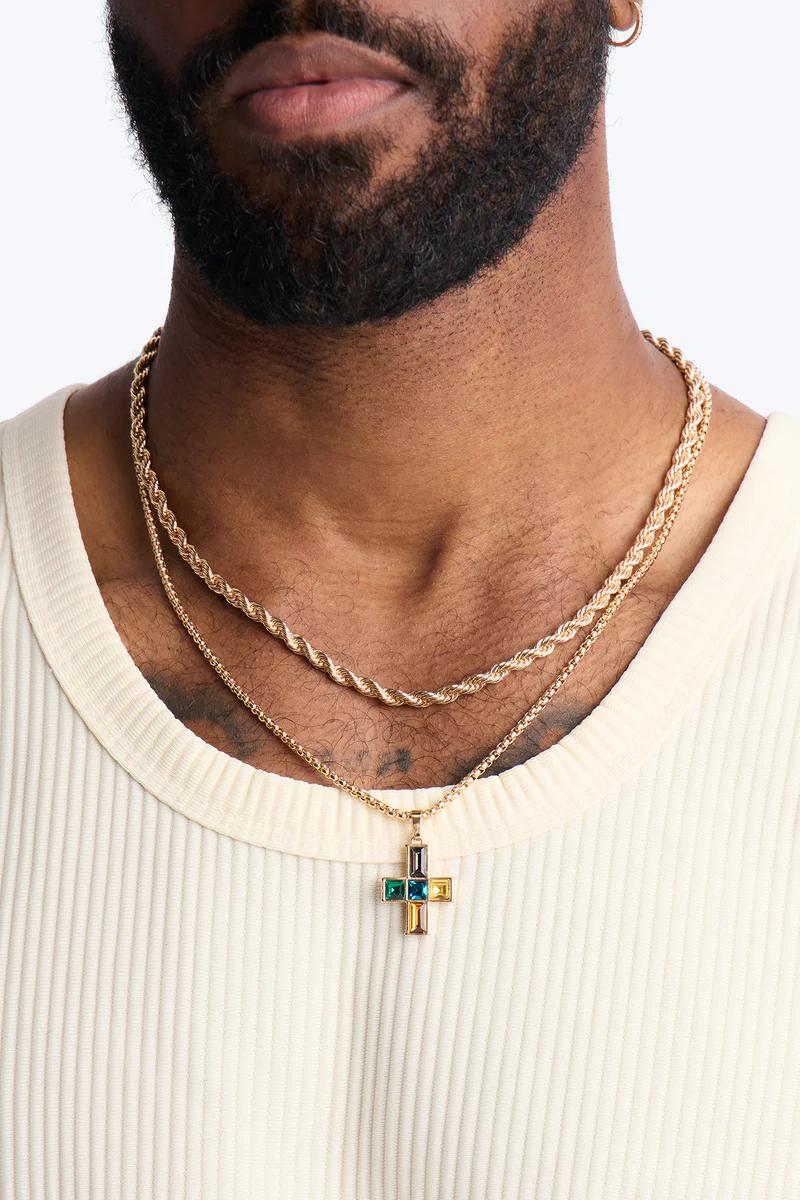 Cadenas de oro en capas, incluyendo una de cuerda, con un colgante de cruz de gemas coloridas en el cuello de un hombre. Exhibiendo joyería masculina.