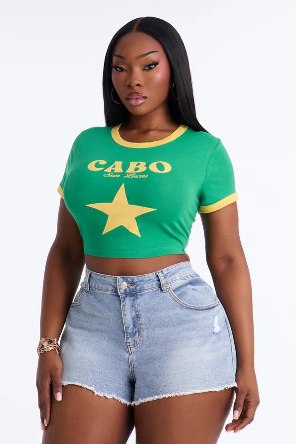 Mujer con camiseta crop verde "CABO San Lucas" amarilla y estrella, shorts vaqueros azules desgastados, joyas doradas.