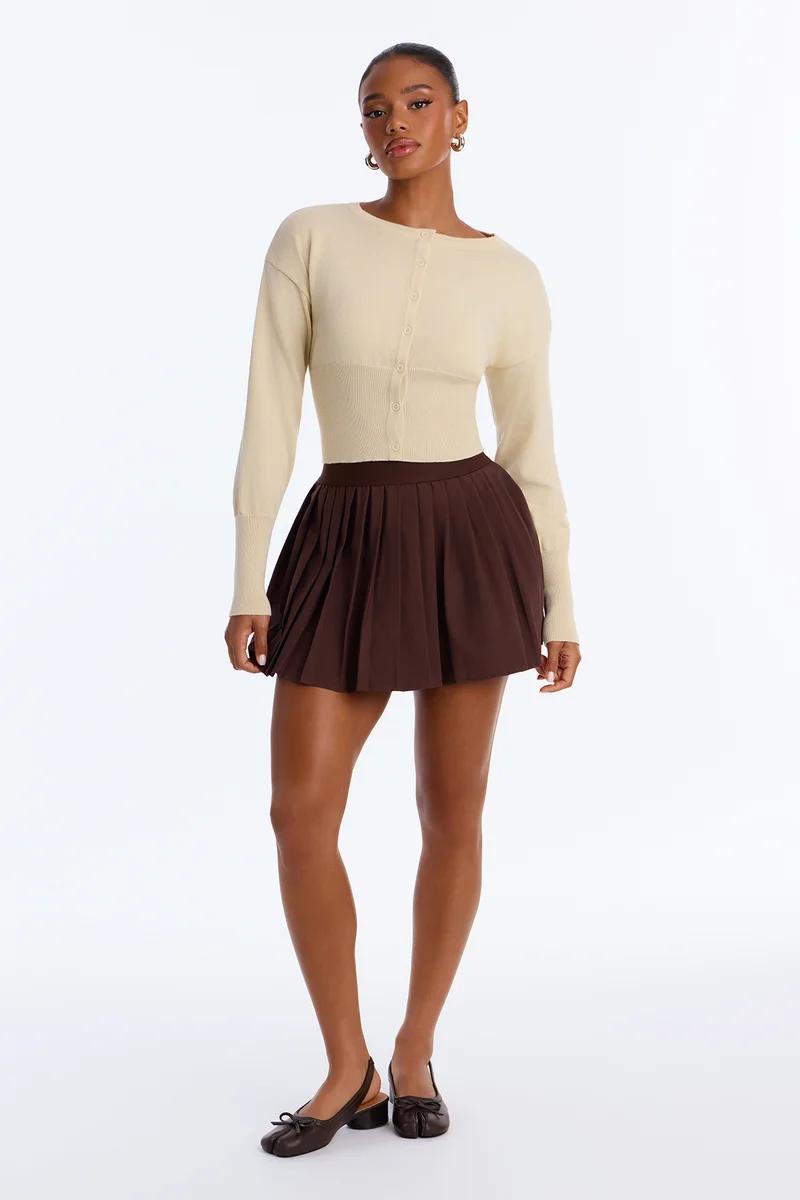 Woman wearing a cream button-up cardigan sweater and brown pleated mini skirt.