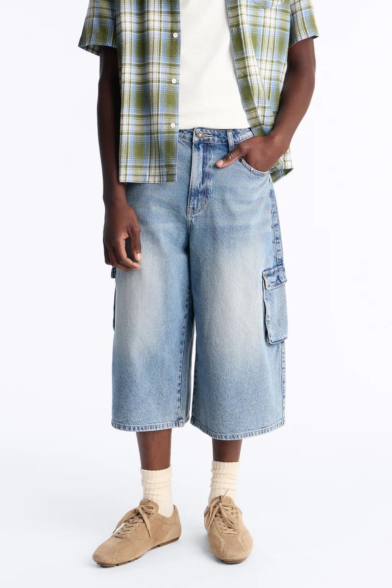 Relaxed men's summer outfit: green/blue plaid shirt, light wash denim cargo shorts with pockets, and tan suede sneakers.