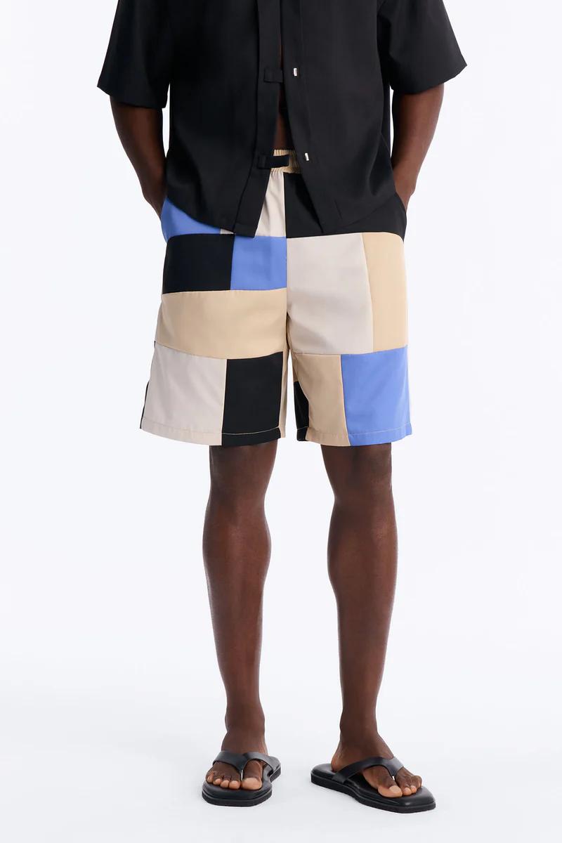Man in black shirt and relaxed patchwork shorts in blue, black, beige, cream, with sandals. Essential summer style.