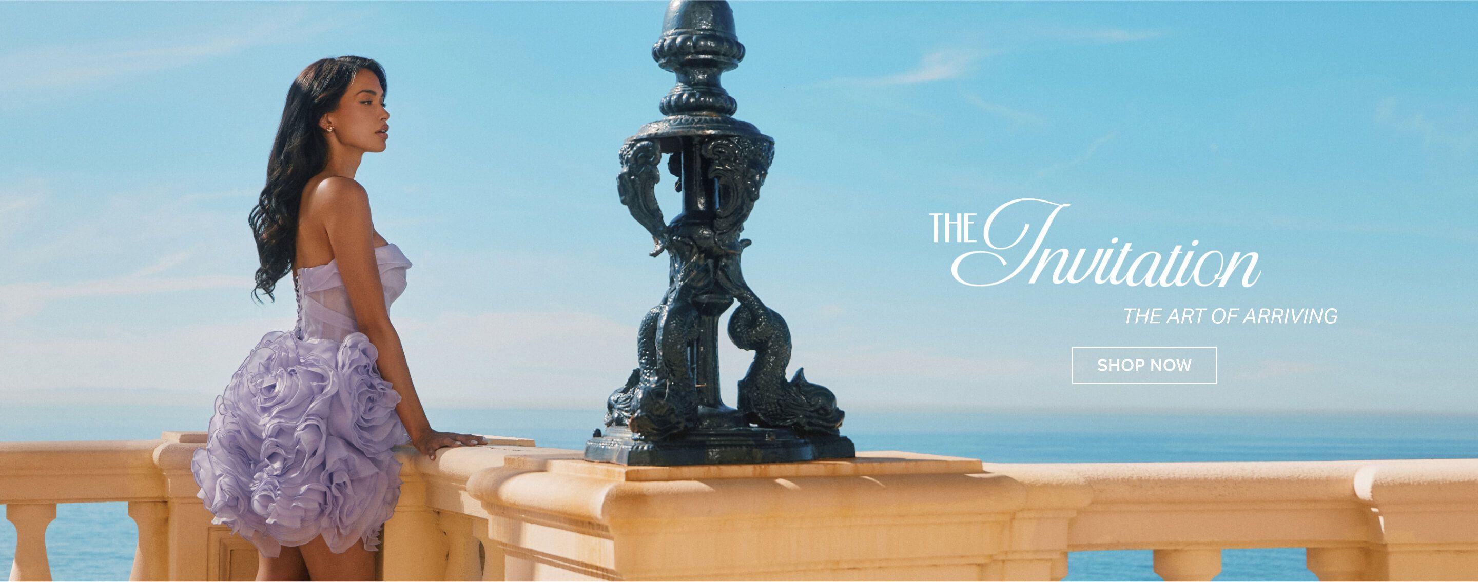 Woman in a flowing lavender strapless dress against a blue sky, relaxing on a railing. Promotes "The Invitation" collection.