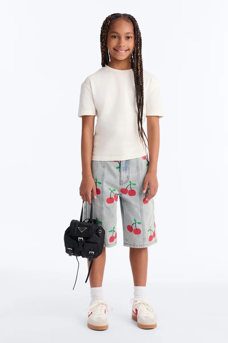 Niña sonriente con camiseta crema, shorts denim de cerezas, zapatillas y mini mochila negra. Moda infantil.