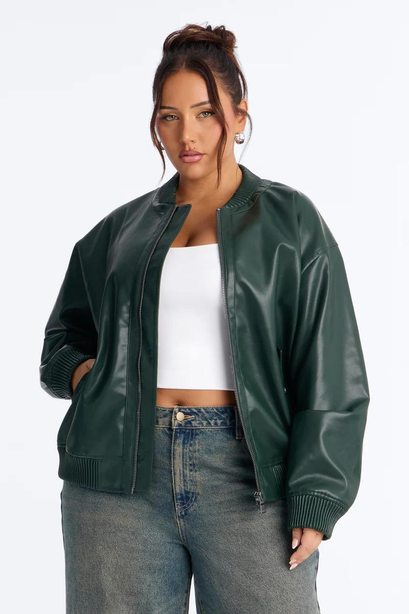 Mujer de talla grande con chaqueta bomber de cuero sintético verde oscuro, top corto blanco y jeans.