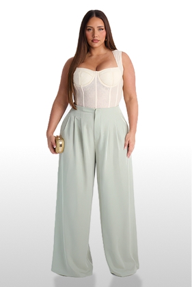Curvy model in cream lace bustier, pleated light green wide-leg pants, holding gold clutch. Highlighting stylish plus-size fashion.