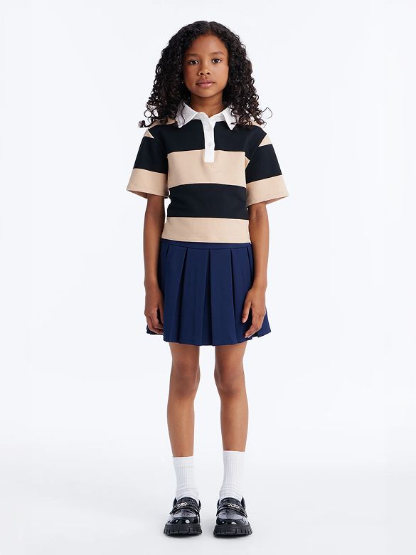 Young girl models a black/beige striped polo, navy pleated skirt, white socks, and black loafers. Stylish kids' collection.