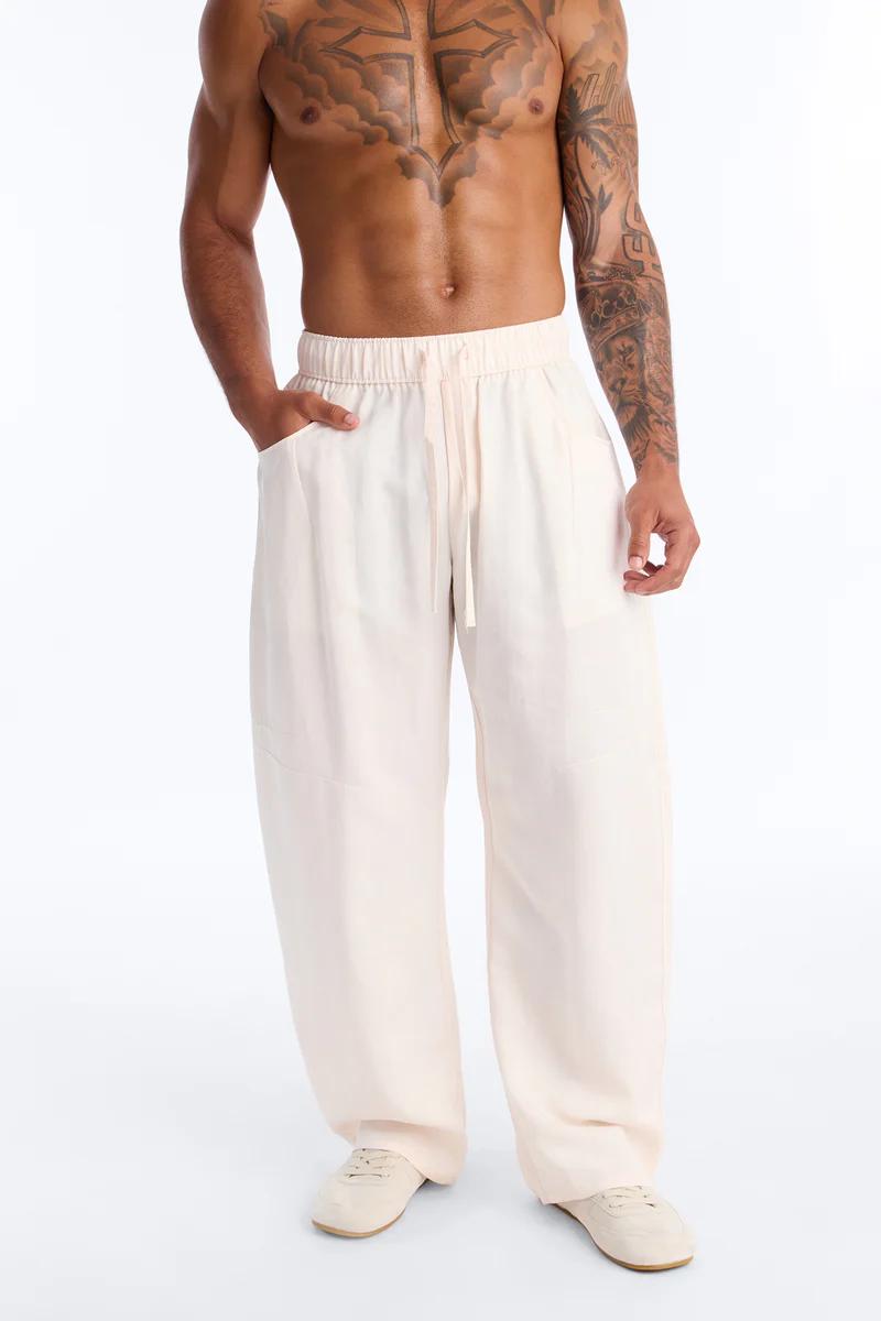 Hombre musculoso modela pantalones de cordón holgados color beige claro con mano en bolsillo, mostrando moda casual masculina cómoda.