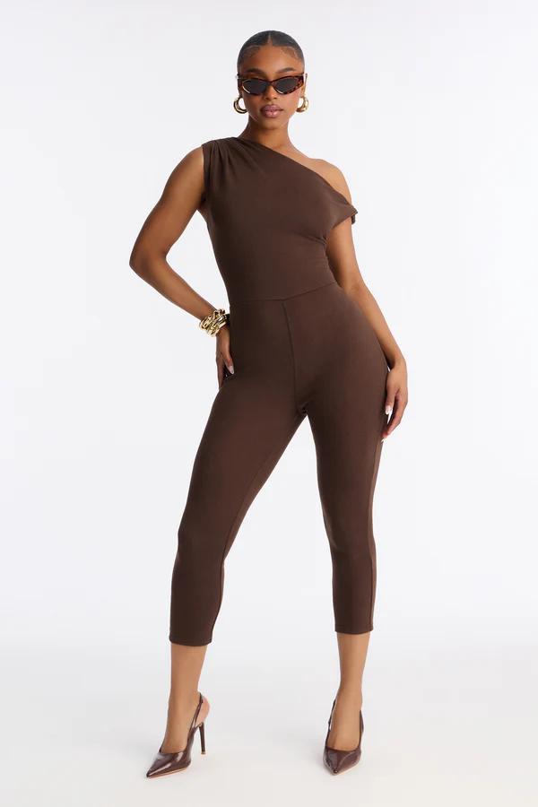 Stylish model in a brown one-shoulder cropped jumpsuit with gold accessories and sunglasses, showcasing chic new apparel.