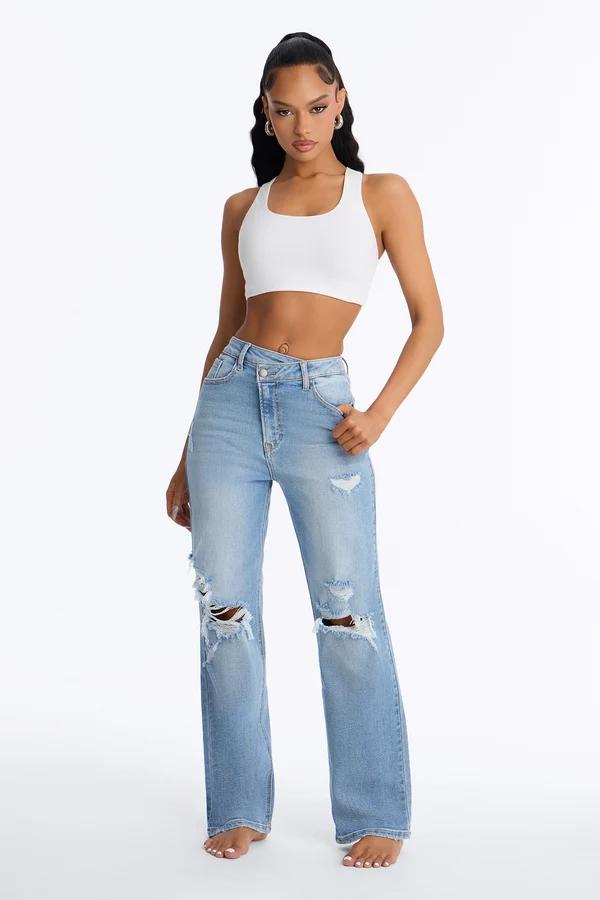 Modelo con jeans rotos de lavado claro, cintura cruzada y top corto blanco, promocionando la nueva colección de denim desgastado.