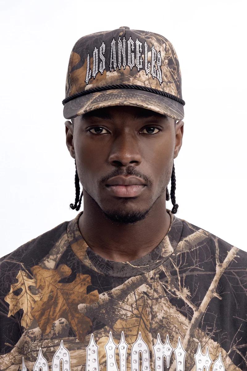 Man wearing a brown camouflage Los Angeles trucker hat with white rhinestones and matching shirt, highlighting urban street style.