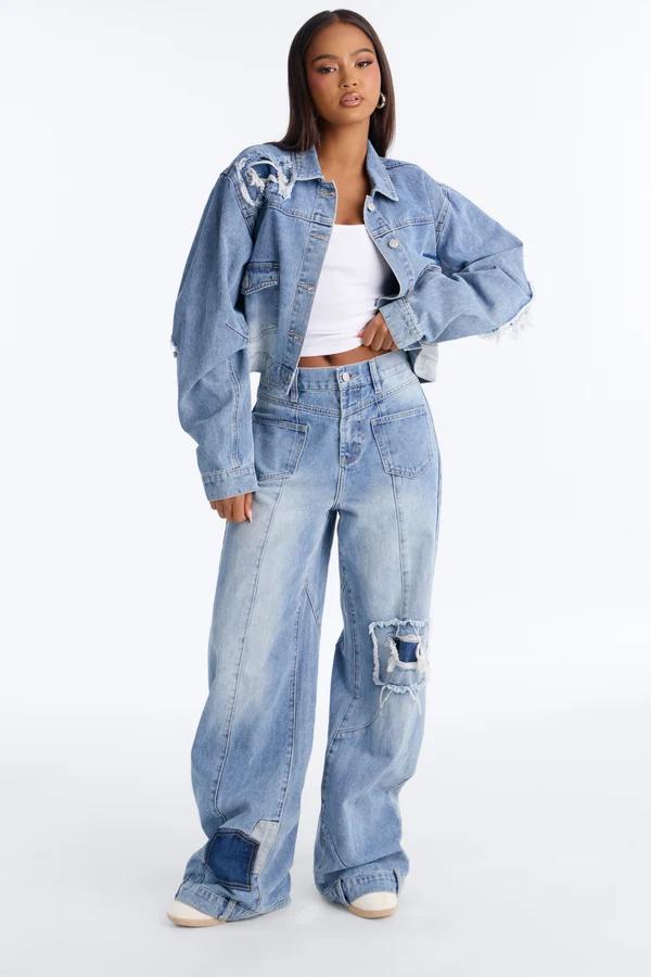 Mujer posa con un conjunto de moda de chaqueta de mezclilla desgastada clara y jeans anchos con un top blanco sobre fondo blanco.