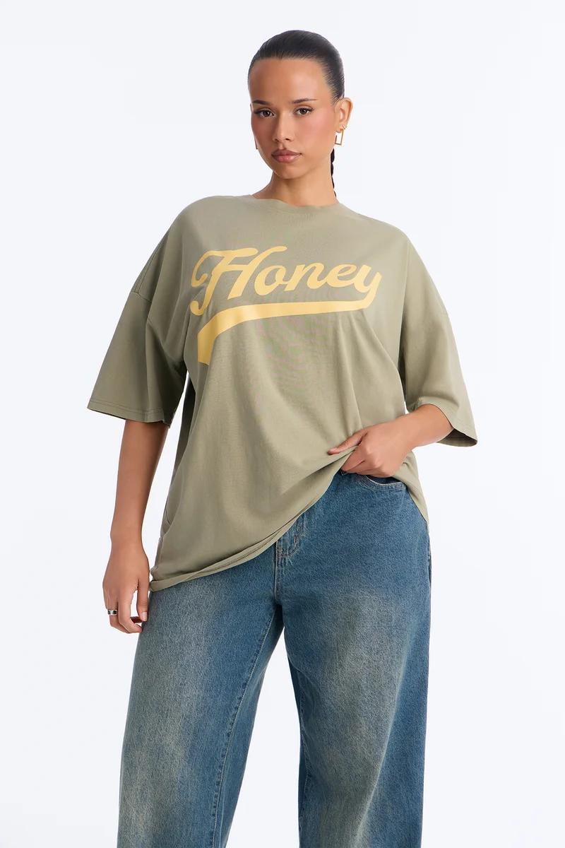 Woman wearing an oversized olive green Honey graphic t-shirt and blue jeans.