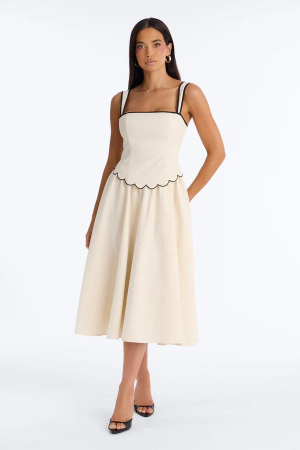 Woman in a square-neck, white maxi dress with blue floral print, bow straps, and a lace hem, holding a straw clutch.