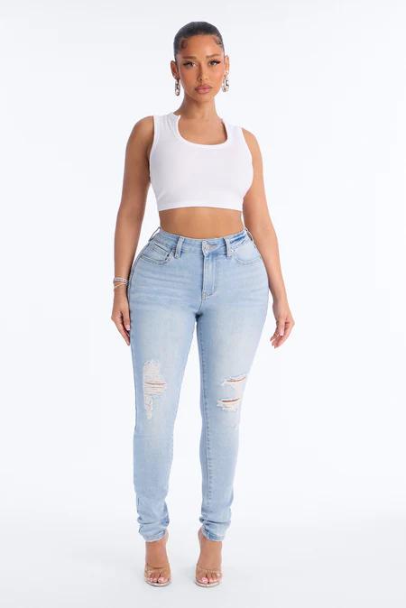 Woman standing with a white sleeveless crop top, light blue ripped high-waisted jeans, and clear heels, on a white background.