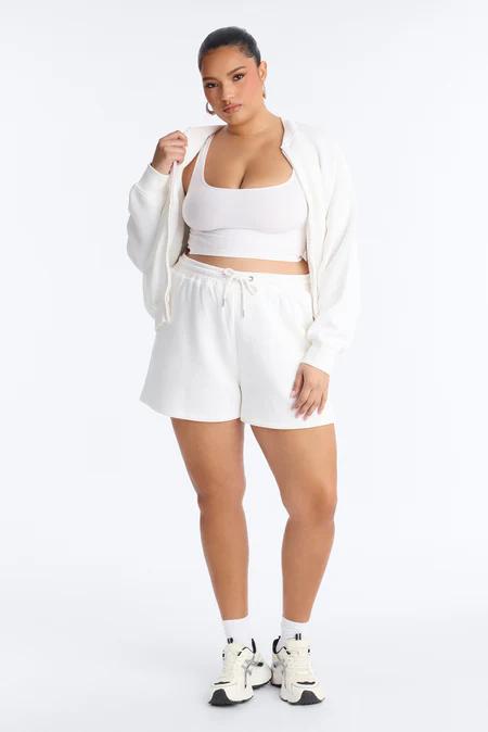 Plus size woman in white athleisure set: crop top, shorts, zip jacket. Comfortable, stylish everyday wear.