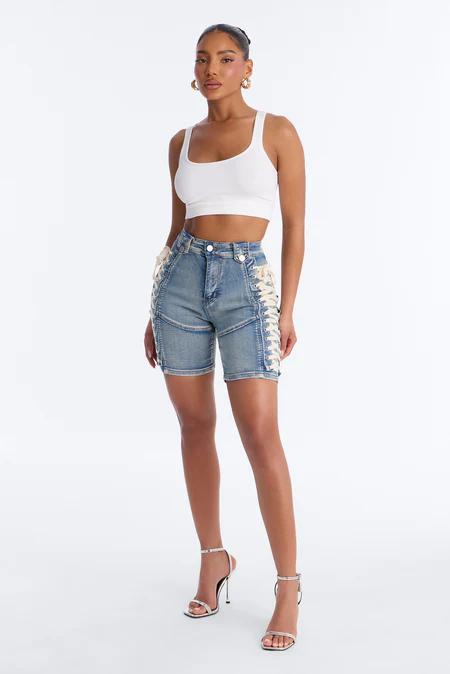 Mujer con top blanco acanalado, shorts ciclistas de mezclilla clara con cordones crema laterales y sandalias de tacón plateadas.