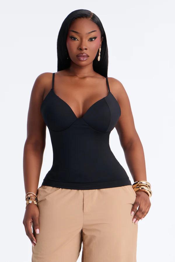 Woman with long black hair wearing a fitted black bustier top, khaki pants, gold dangle earrings, and bracelets. White background.