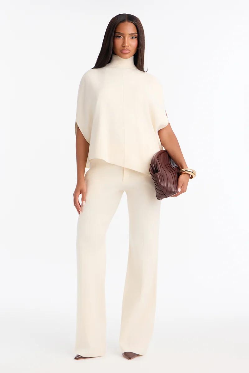 Woman wears an elegant cream turtleneck top and matching wide-leg trousers paired with a brown clutch, showcasing a chic fashion collection.