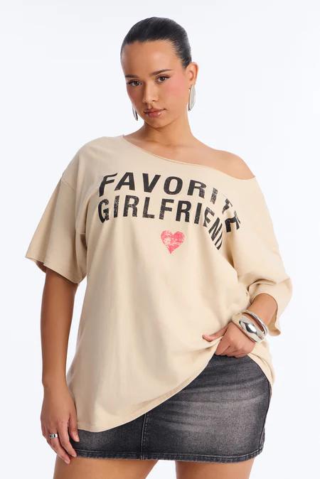 Mujer con camiseta gráfica beige hombro descubierto "Favorite Girlfriend" con corazón rojo y falda vaquera negra. Moda casual.