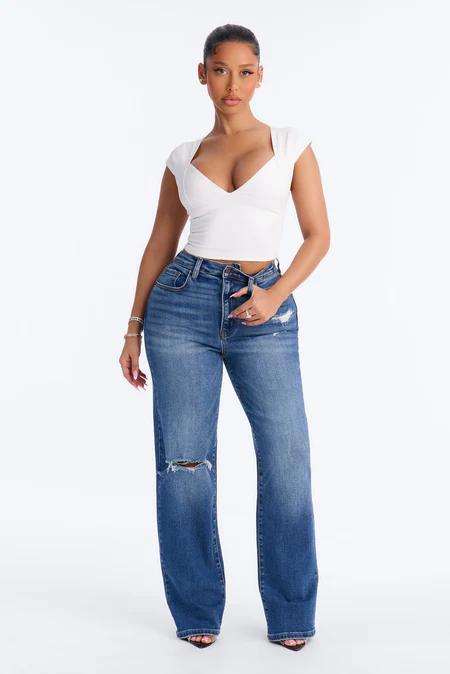 Woman wearing a white sweetheart-neck crop top and distressed wide-leg blue jeans, confidently posing against a seamless white background.