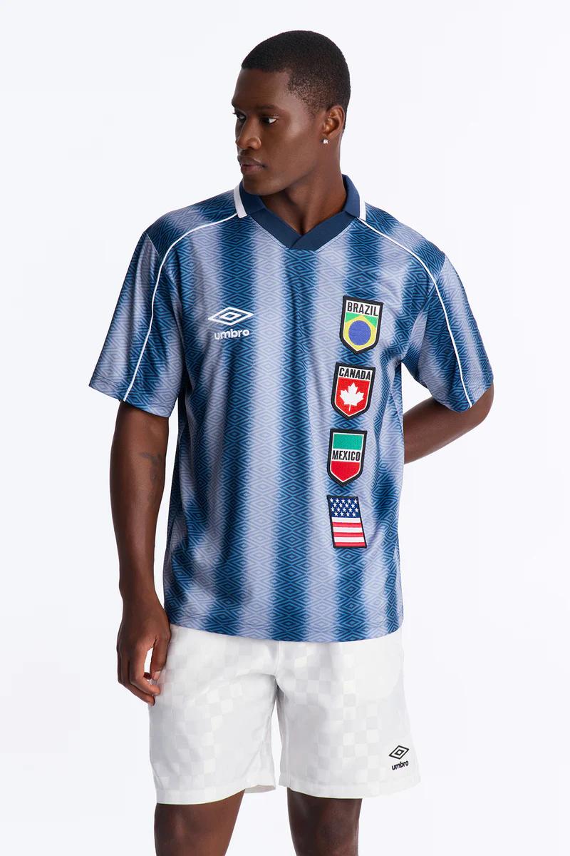 Hombre modela camiseta Umbro azul estampada con banderas internacionales y shorts blancos, mostrando nueva colección deportiva.