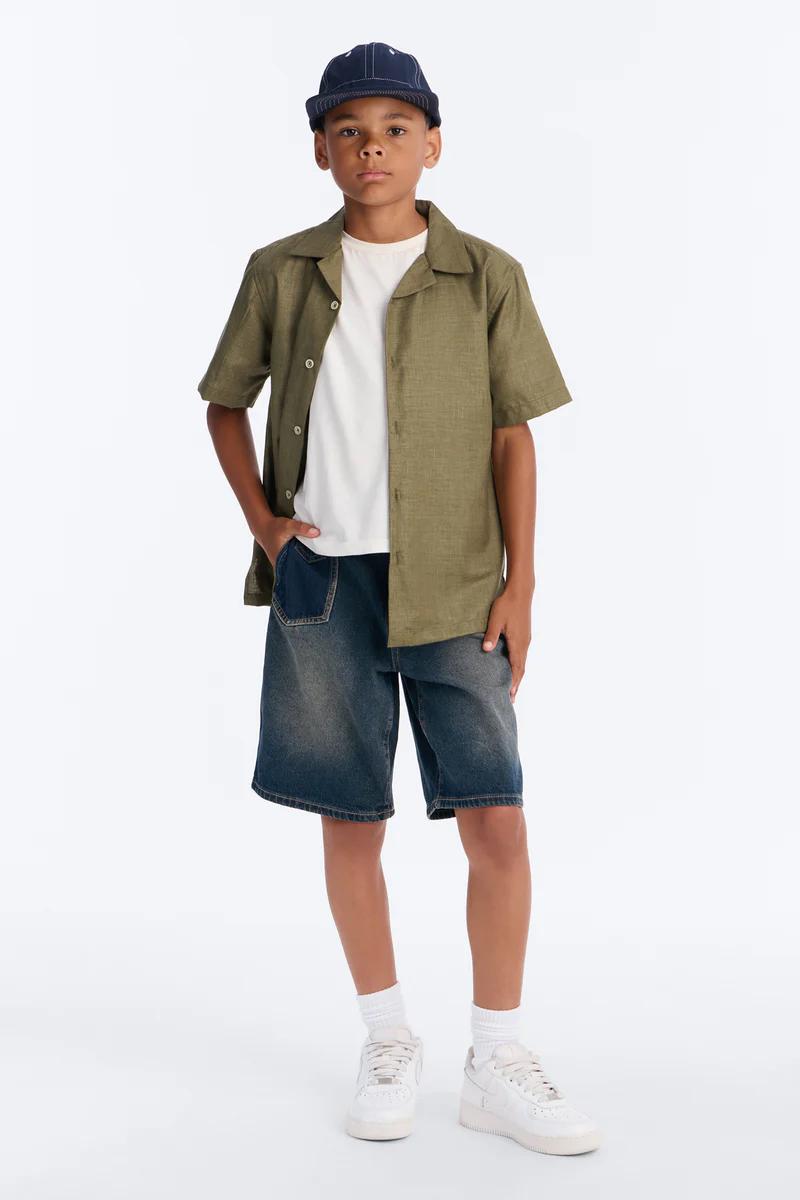 Niño con gorra azul, camisa verde abierta, camiseta blanca, shorts mezclilla, tenis blancos.
