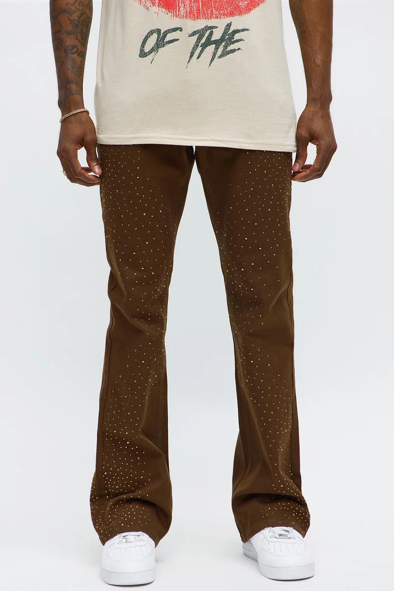 Man in stylish brown studded pants with gold detailing, white sneakers, and a beige graphic tee, highlighting new men's fashion.
