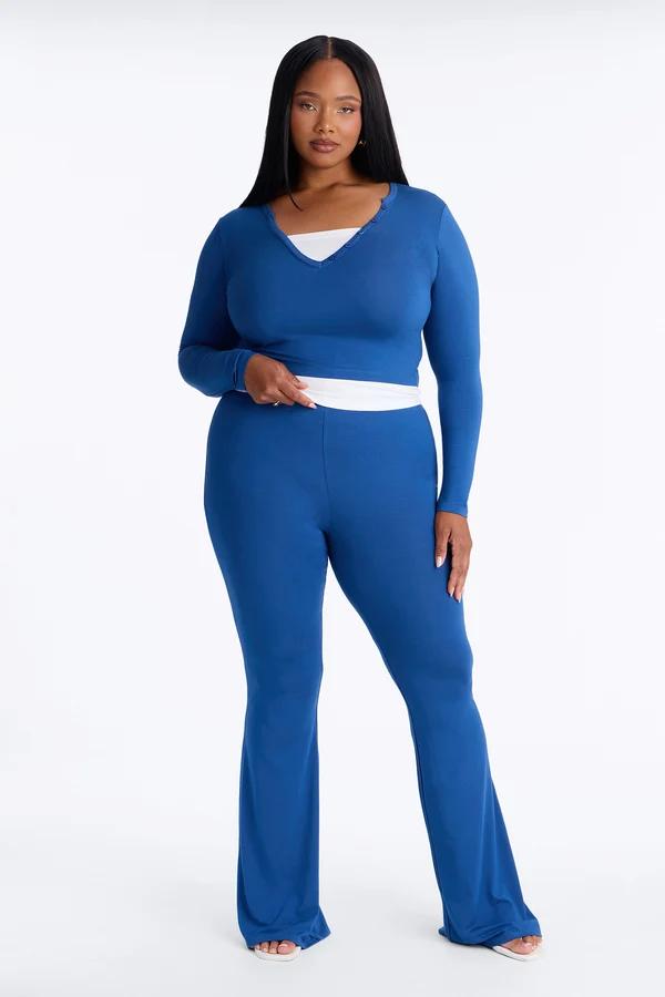 Plus size model wears a blue long-sleeve crop top and matching flared pants from a new cozy loungewear collection against a white background.