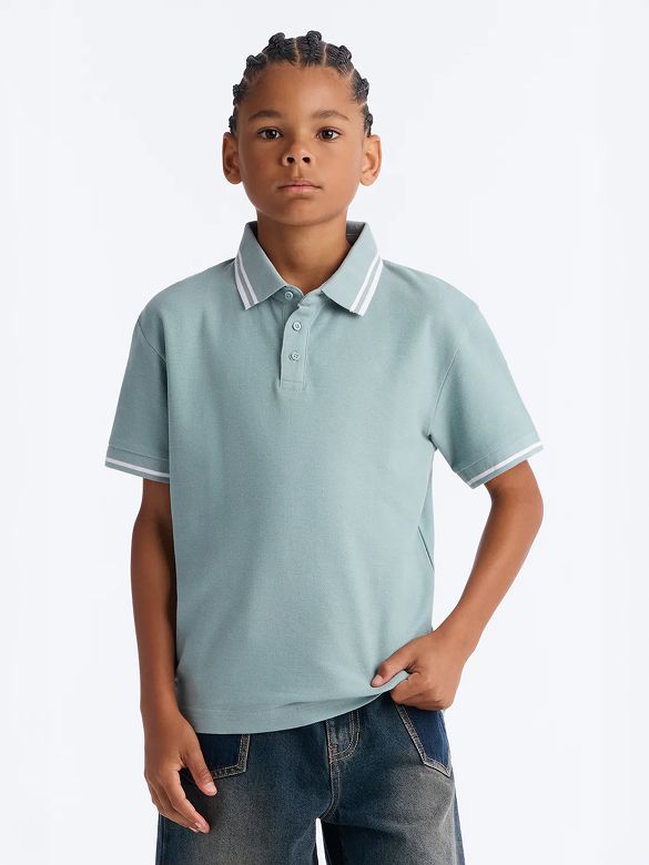 Boy modeling a dusty blue short-sleeve boys' polo shirt with white striped collar and cuffs, paired with blue jeans. Shop kids' casual clothing.