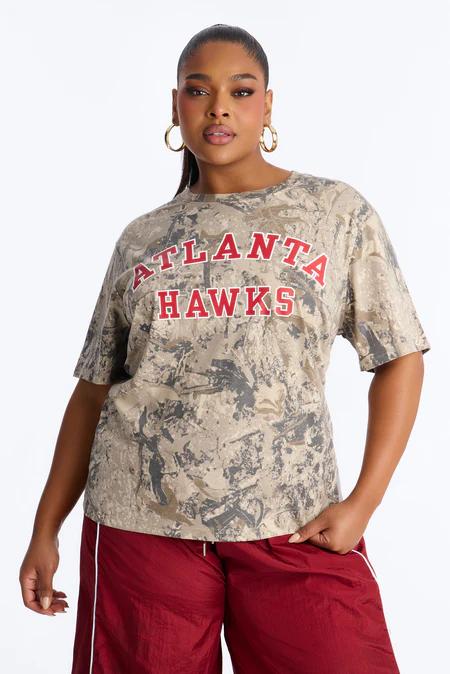 Mujer segura con camiseta beige camuflada de Atlanta Hawks con letras rojas y pantalones cortos rojos, promoviendo moda casual talla grande.