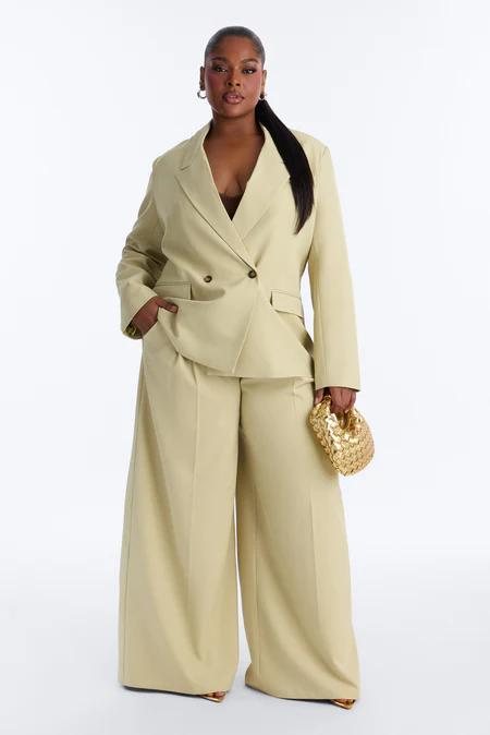 Mujer con blazer cruzado y pantalones anchos verde oliva claro, llevando un bolso dorado trenzado. Cabello oscuro en coleta pulcra.