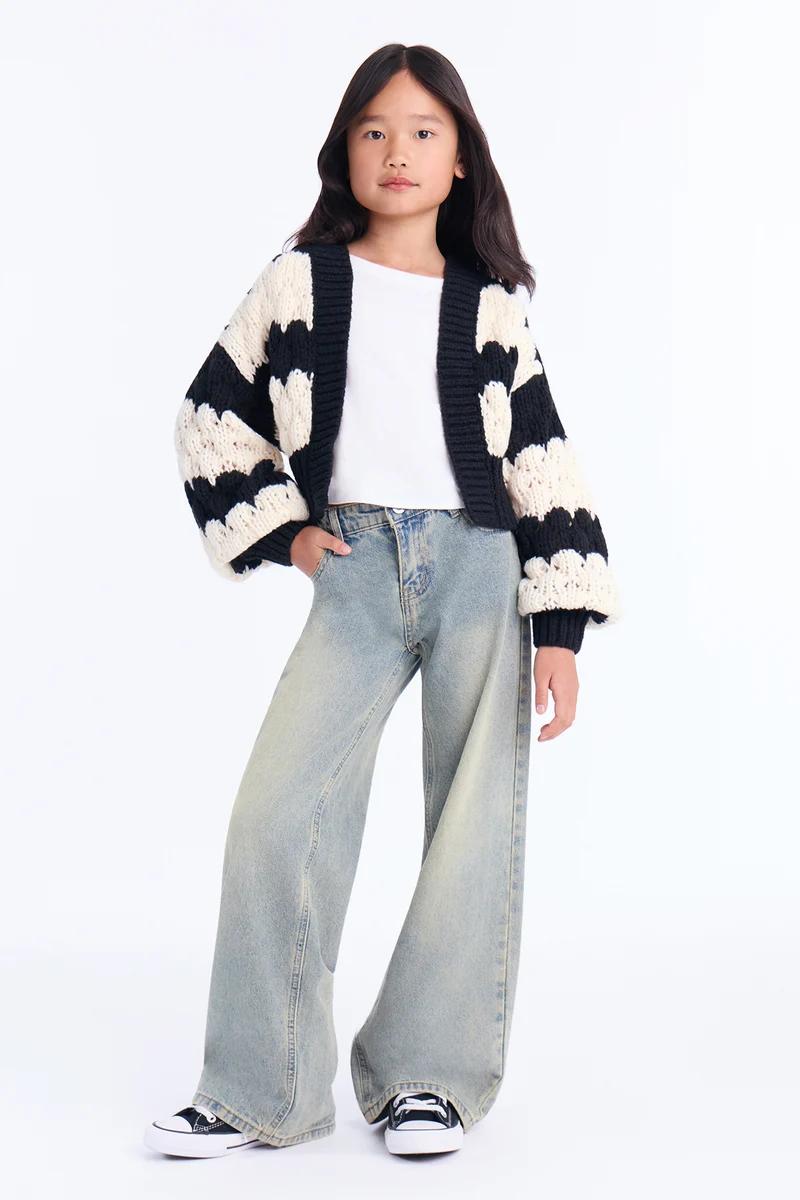 Niña con cárdigan de punto grueso en blanco y negro, camiseta blanca y vaqueros de pata ancha.
