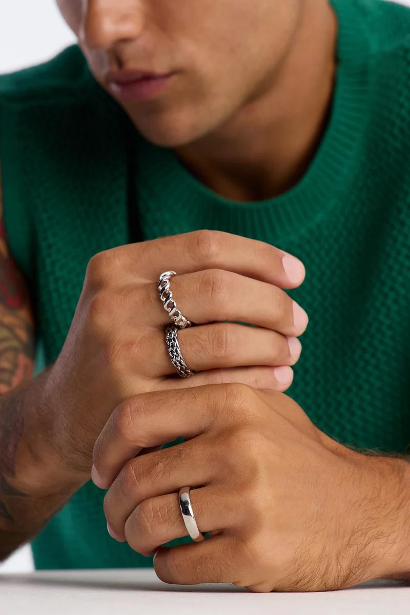 Primer plano de manos de hombre con tres anillos de plata: banda lisa, eslabón cubano y diseño trenzado. Destacando joyas masculinas de moda.