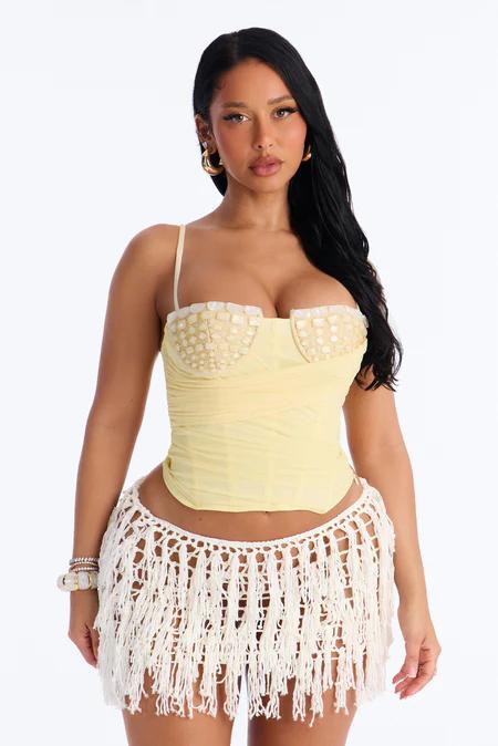 Mujer con corsé amarillo y copas blancas adornadas, falda blanca de ganchillo con flecos, aros dorados y pulseras.