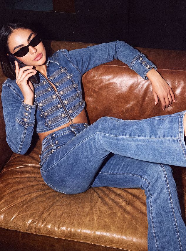 Woman in cropped denim jacket & flared jeans with metal details, sitting on a brown couch, talking on phone. Discover new denim styles.