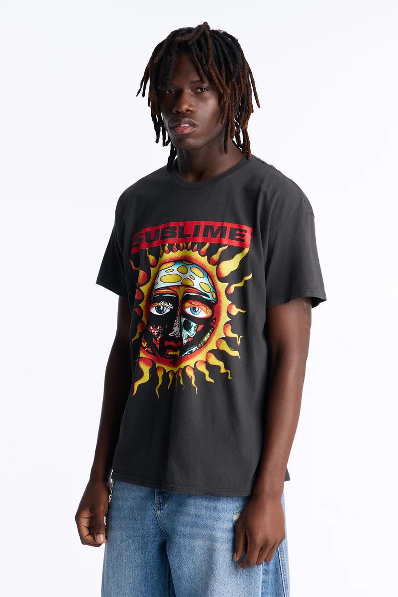 Man wearing a dark grey Sublime band t-shirt featuring a vibrant sun graphic, styled with blue jeans. Shop men's graphic tees.