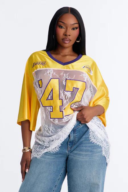 Persona con una camiseta de Los Lakers amarilla y morada, con paneles de encaje blanco transparente, el "47" y texto, y jeans azules.